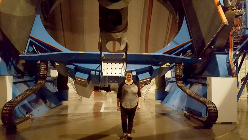 Astrophycist Mallory Molina is standing in front of a huge telescope. The telescope has a blue base with a long silver body.
