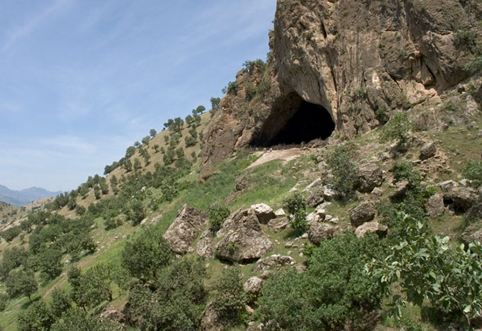 A photo of a rocky scrubby hill. At the top there is a steep cliff with a very large cave opening.