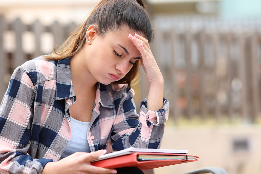 A young white woman wearing a pink and black plaid shirt is sitting outside with her forehead on her hand. Her other hand holds a binder and some notebooks. She's puffing her cheeks out and appears stressed out.